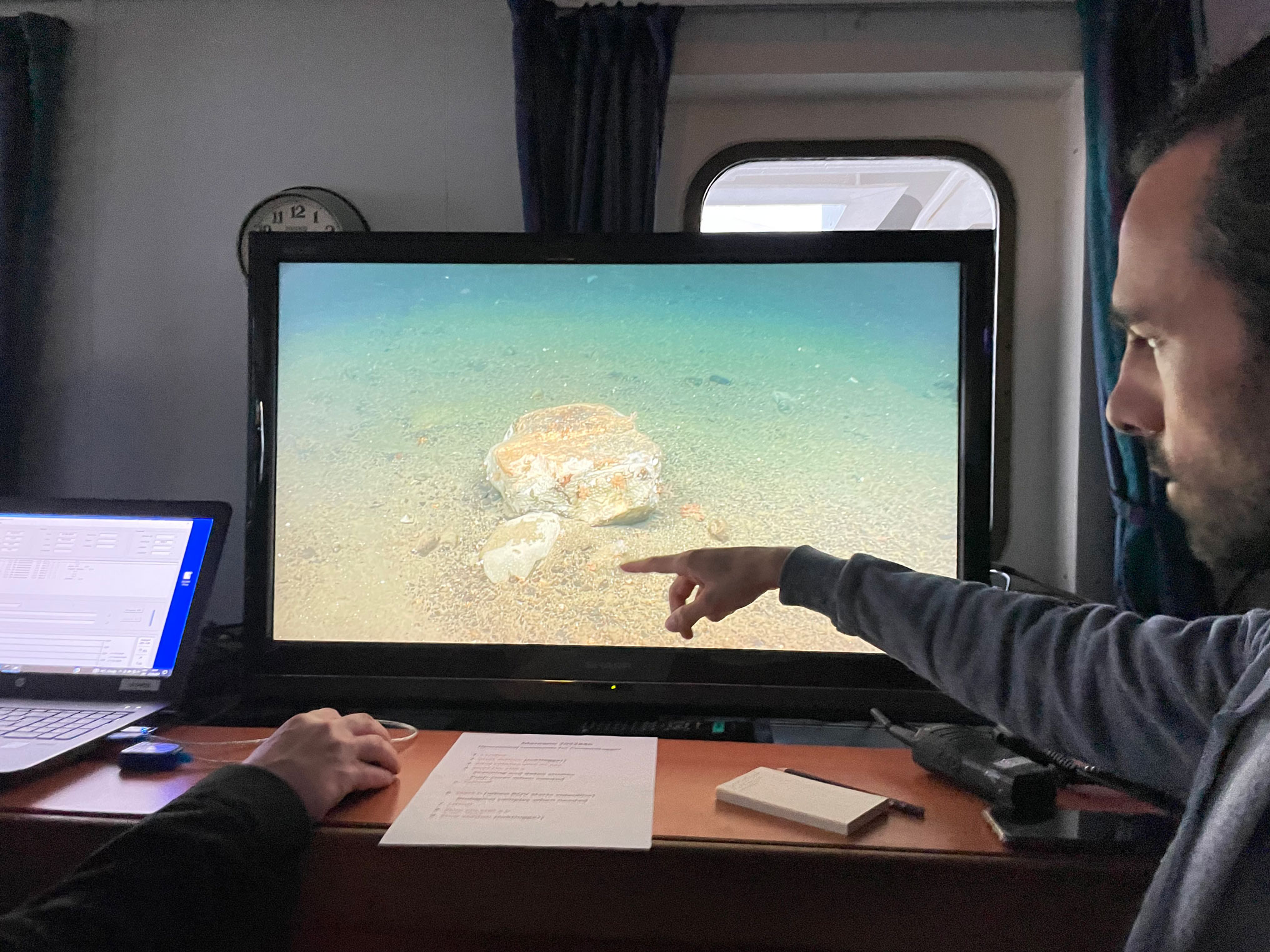 Bilde av forsker som peker på en stein som har dukket opp på tv-skjermen som viser bildene fra havbunnen.