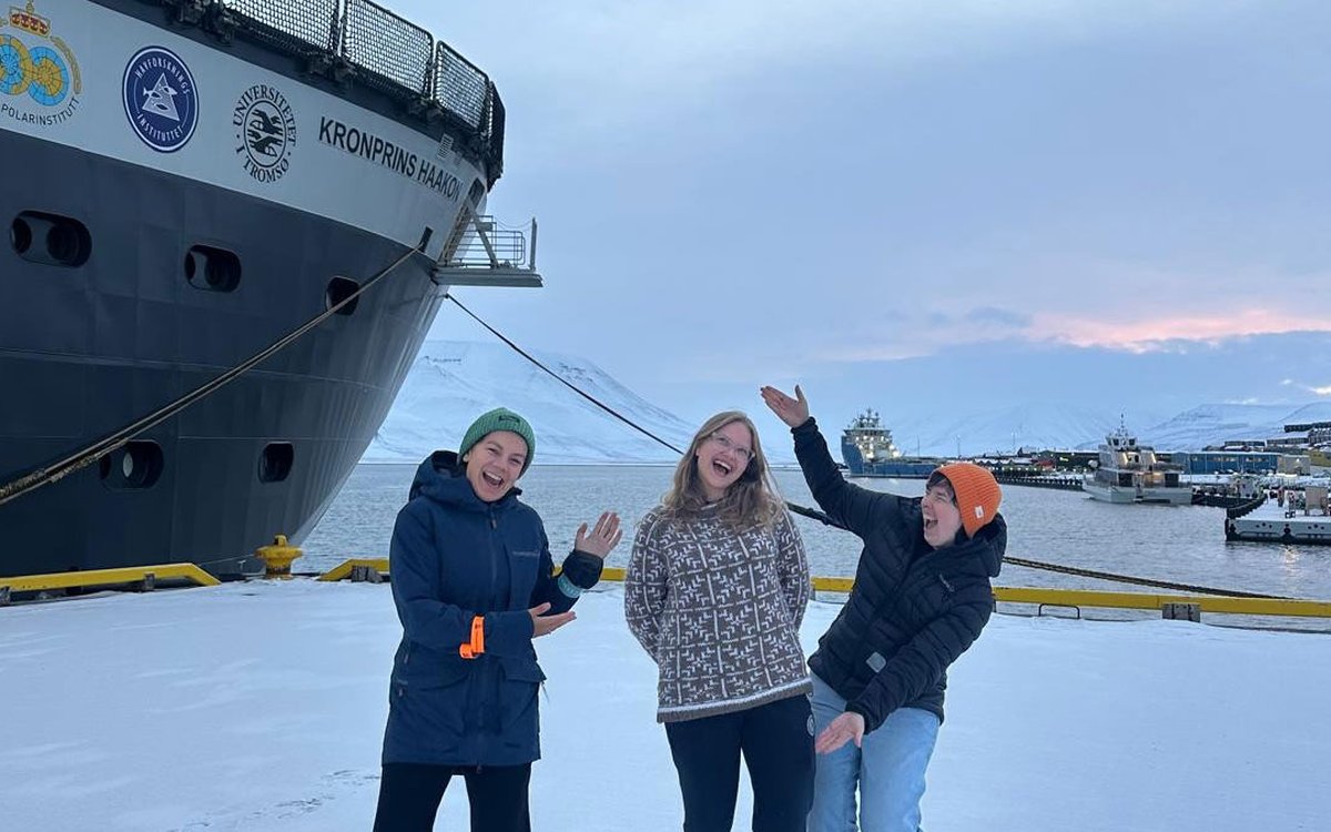 Tre blide og jublende personer står på en snøket kai. En stor båt ligger fortøyd. I bakgrunnen hav og himmel.