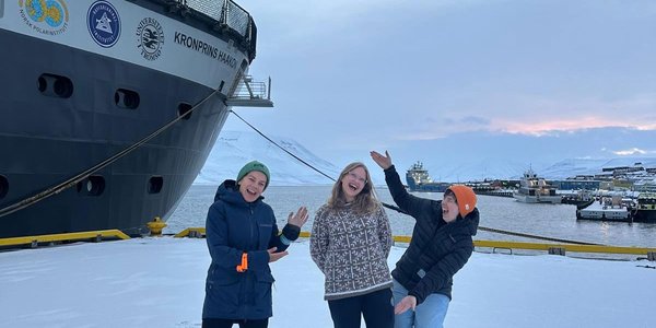 Tre blide og jublende personer står på en snøket kai. En stor båt ligger fortøyd. I bakgrunnen hav og himmel.