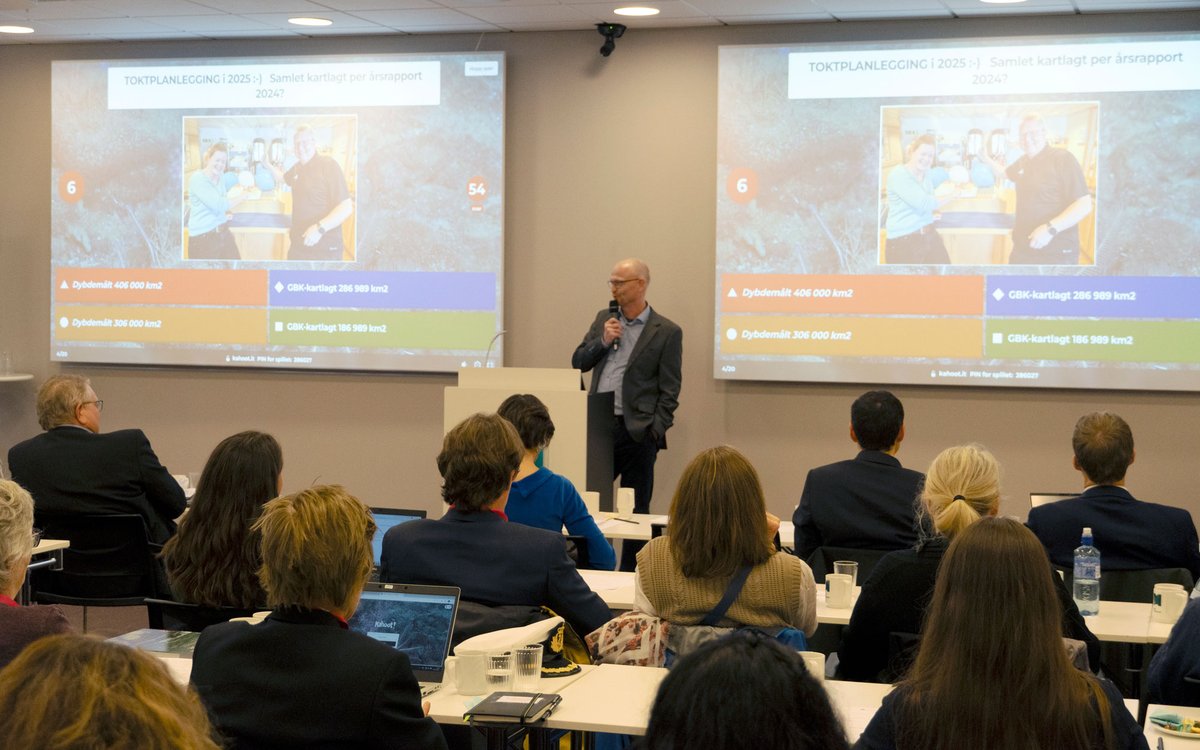 Ein mann står foran publikum og snakkar i mikrofon. Bak han på veggen er det skjermar med spørsmål og svaralternativ.