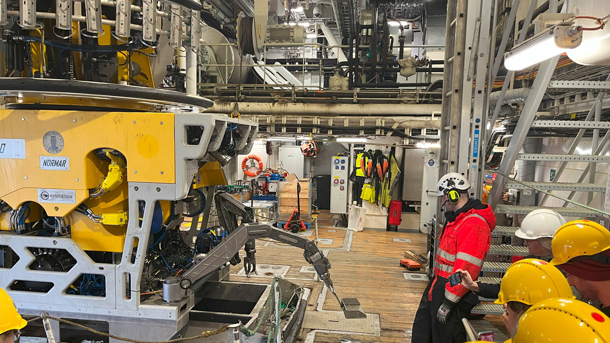Inne i en slags industrihall på forskningsfartøyet. Tredekk. En stor gjenstand står på gulvet. Den har ulike redskaper. Rundt står personer og ser på den. 