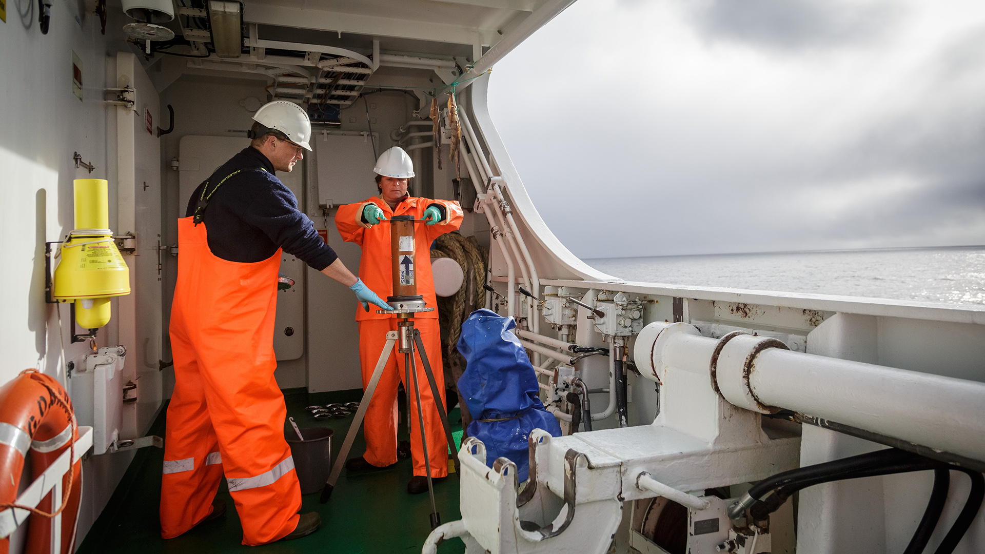 En mann og en kvinne står på dekket på en båt og holder en kjerneprøve på et stativ mellom seg. De deler prøven opp i tynnere skiver. På siden av bildet er det utsikt utover havet. 