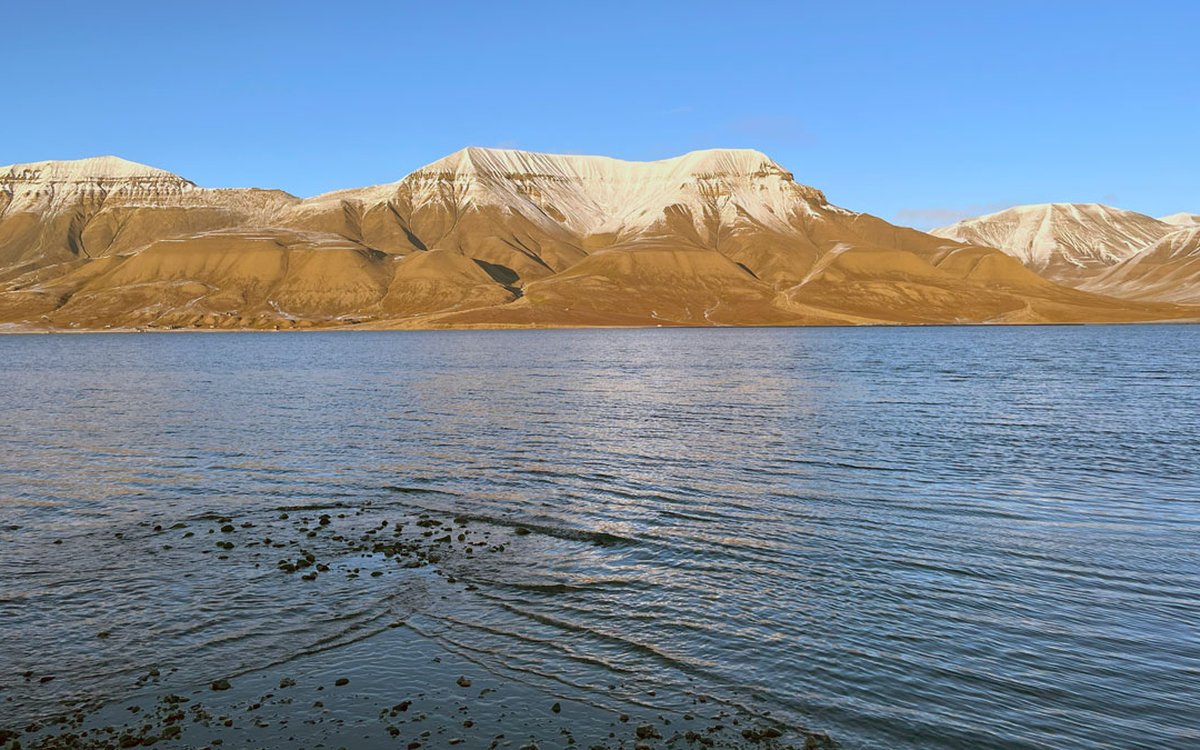Landskap. Sjø i forgrunnen, høye og bratte fjell med snø på toppen og blå himmel.