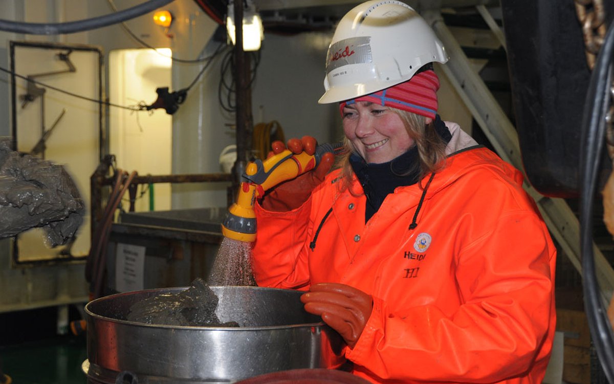 En kvinne med hjelm, hansker og oransje regnklær spyler oppi en metallbeholder med en vannslange. HUn smiler.