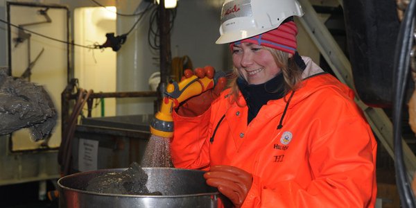 En kvinne med hjelm, hansker og oransje regnklær spyler oppi en metallbeholder med en vannslange. HUn smiler.