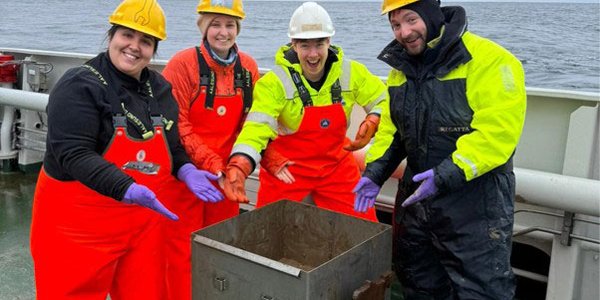 Fire personer står glade rundt en metallkasse. De smiler og peker på kassen.