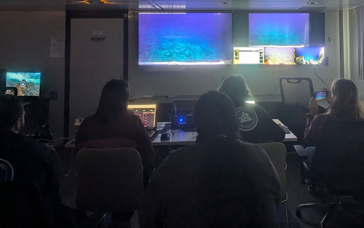 Fire personer sitter i et mørkt rom og ser på to store og flere små skjermer.