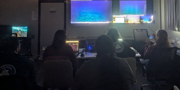 Fire personer sitter i et mørkt rom og ser på to store og flere små skjermer.